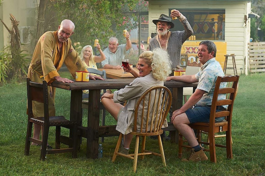 Two women and four men sit around two tables in a smokey backyard drinking four x branded beers