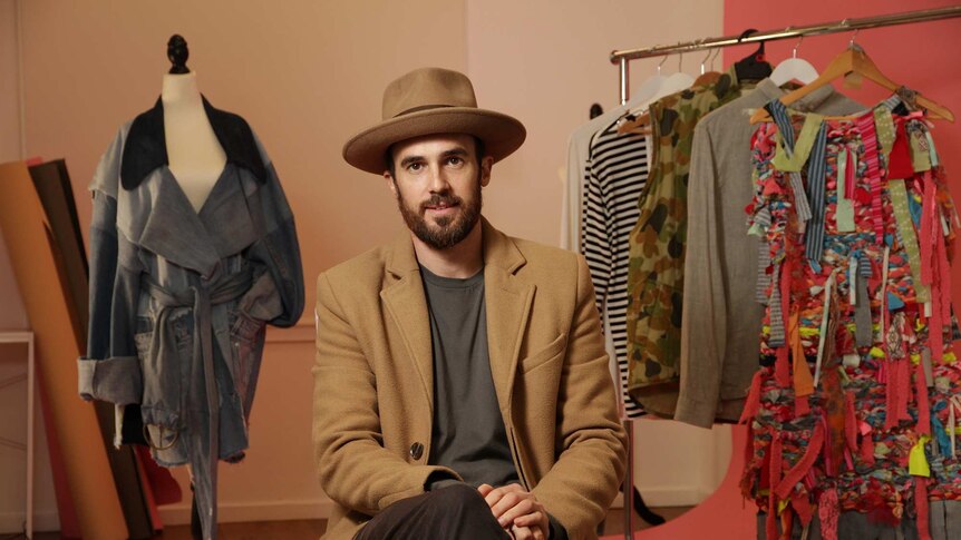 A well dressed man in a hat and a coat surrounded by clothing in a fashion and photography studio