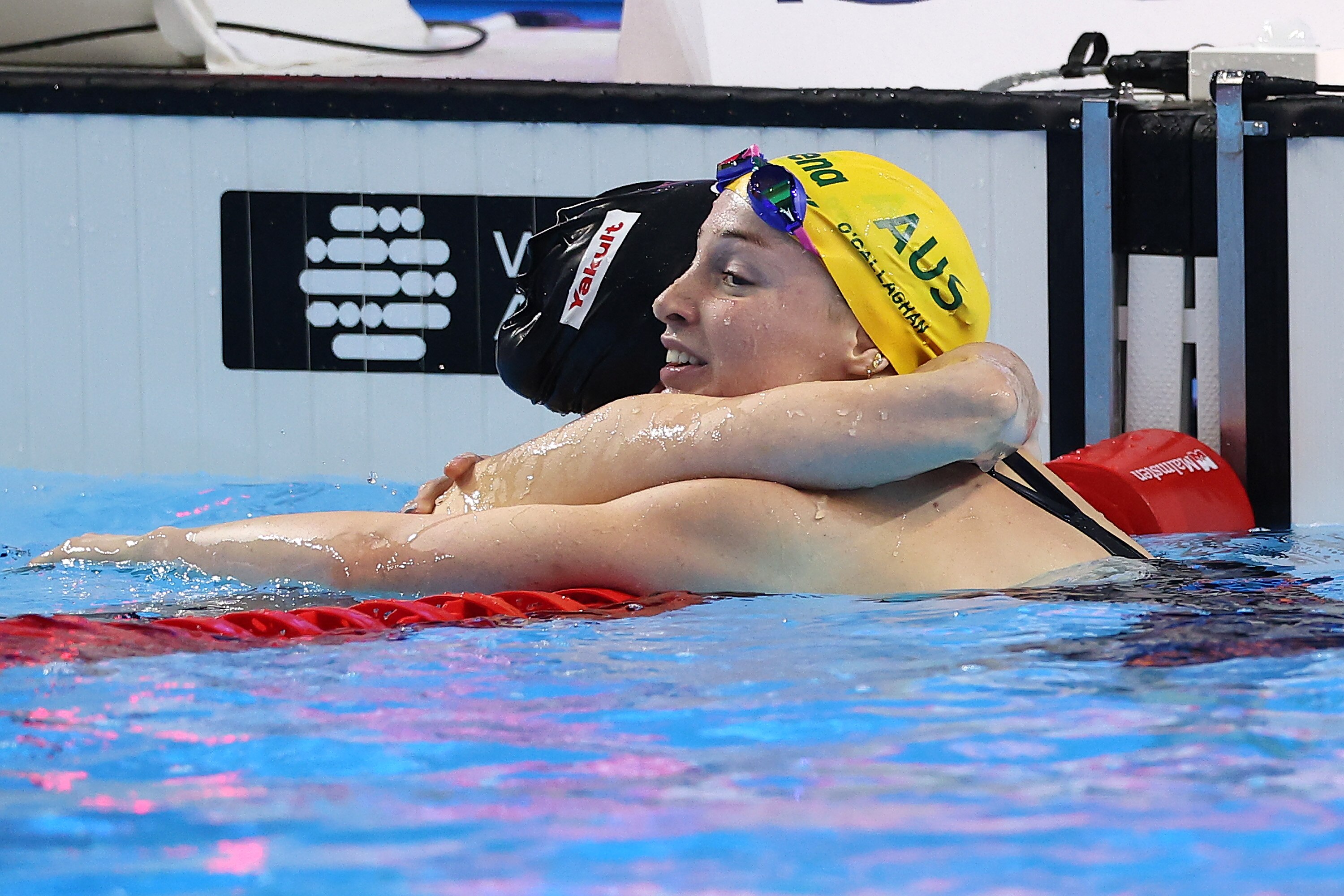 Two swimmers, one in yellow cap and one in black embrace at the end of a pool.