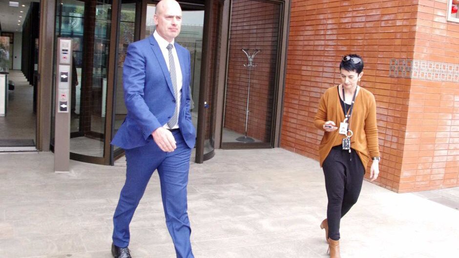 Dean Nalder walks out of a building wearing a blue suit with a woman nearby.