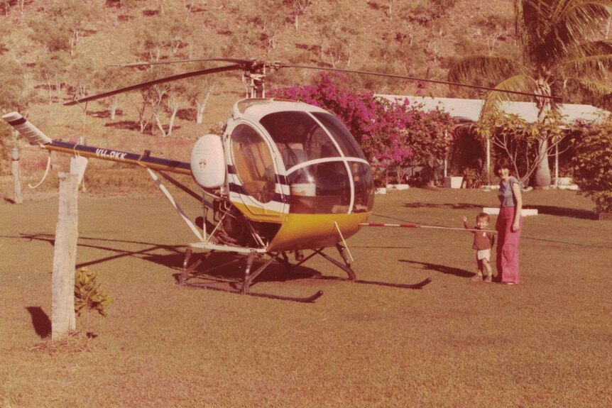 An old photograph of a small mustering helicopter. Polly Kim and one of her young children stands beside it.
