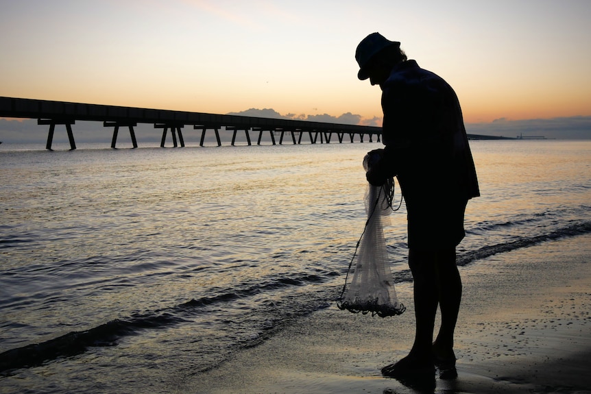 A silhouette of a man holding a fishing net on the edge of the water with a long jetty in the background.