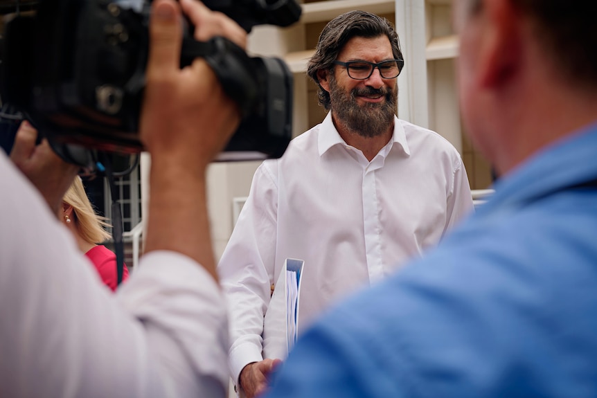 A man wearing a white shirt with a dark beard walks past TV cameras.