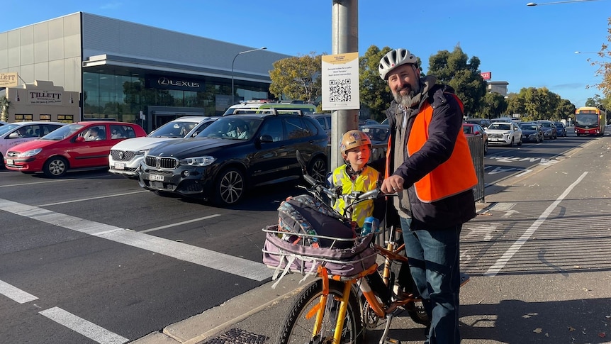 man and boy with bike helmets and hi-vis vests standing next to bicycle at an intersection cars in background