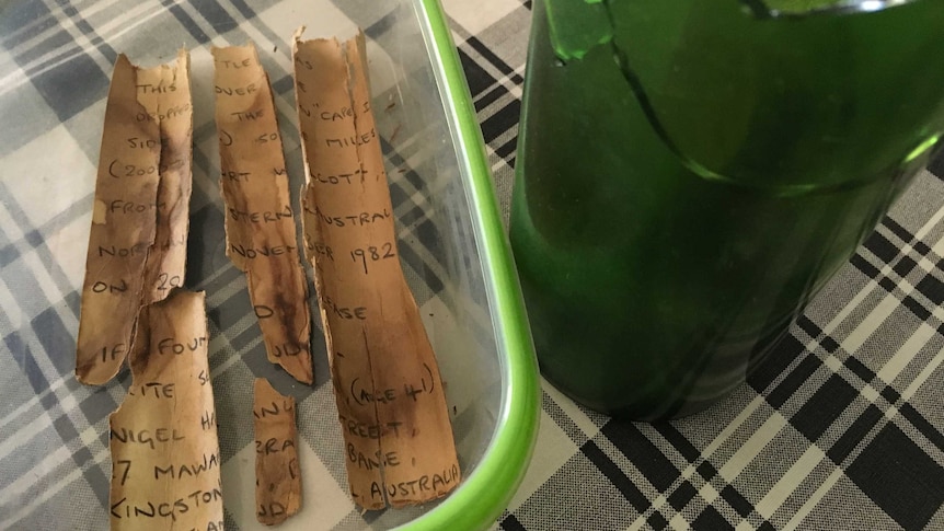 A smashed green bottle and a broken letter in a clear container sit on a table cloth