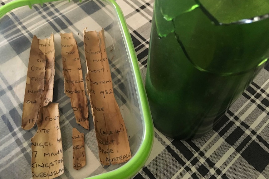 A smashed green bottle and a broken letter in a clear container sit on a table cloth