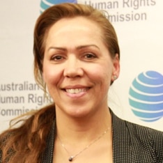 Long headshot of a woman with long hair, pulled to one side, standing in front of Australian Human Rights Commission poster