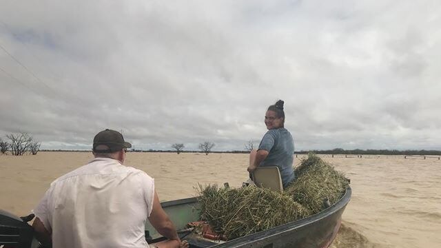 A woman and a man travel sitting on a boat loaded with hay, moving through brown flood waters.