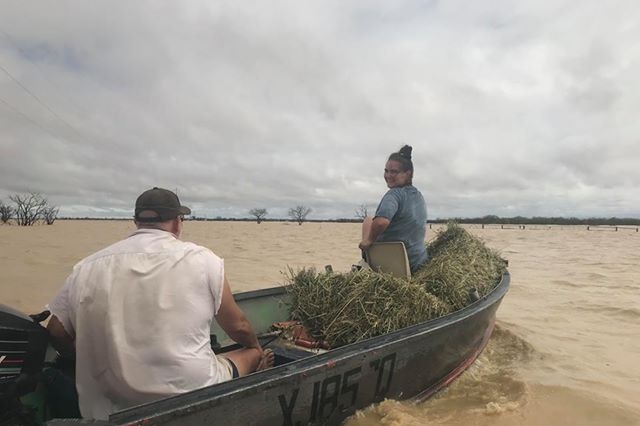 A woman and a man travel sitting on a boat loaded with hay, moving through brown flood waters.