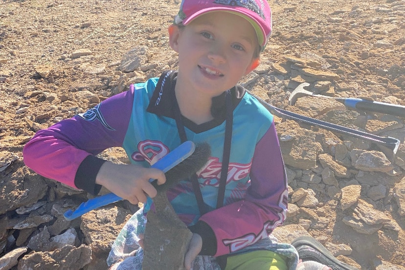 A young girl sits on the ground and holds up a fossil. 