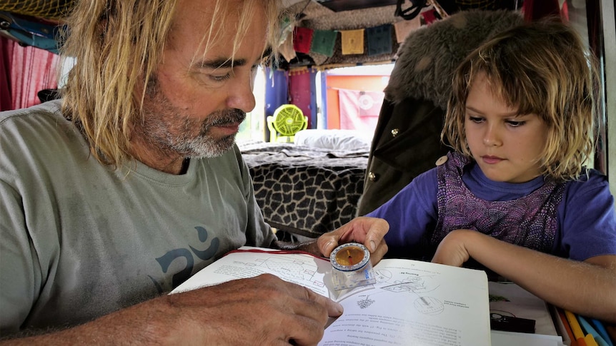 Father and daughter sitting around a small table in a campervan-style bus with a compass and an open book.