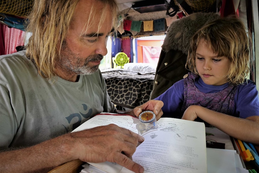 Father and daughter sitting around a small table in a campervan-style bus with a compass and an open book.