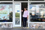 A woman with long black hair and wearing a pink top stands in the door of a supermarket