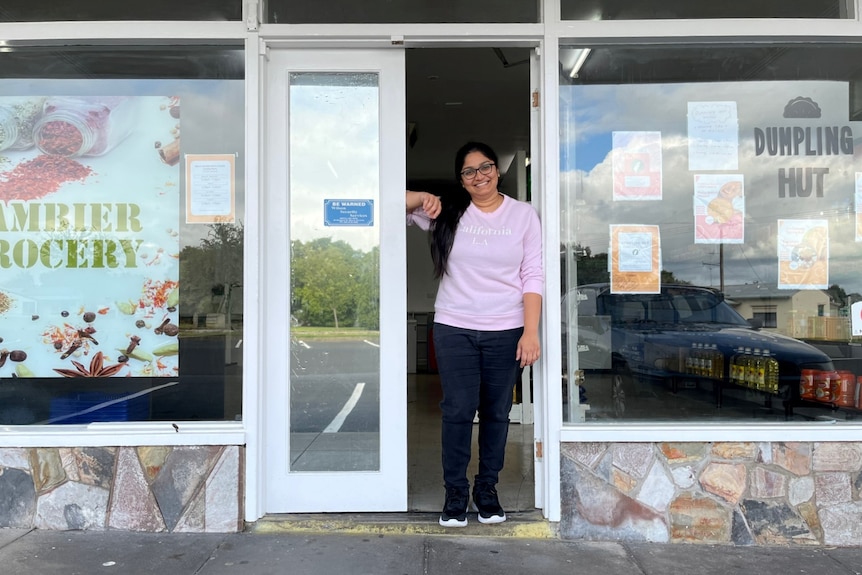 A woman with long black hair and wearing a pink top stands in the door of a supermarket