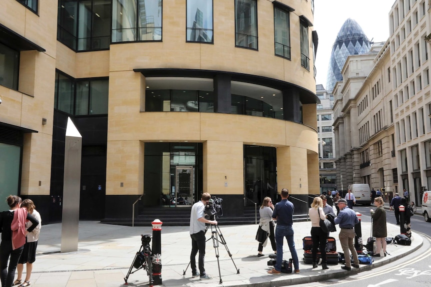 A throng of journalists stand on the pavement outside a curved sandstone building on a narrow London street.
