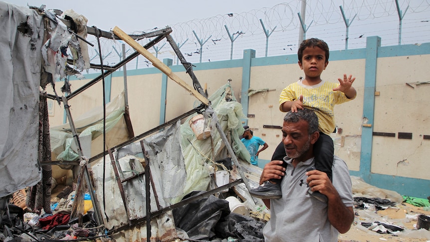 A child sitting on the shoulders of a man looking confused and raising his arm. A backdrop destroyed tents and rubble
