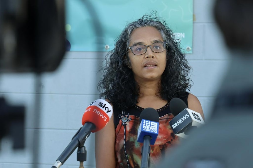 A woman standing in front of news microphones, seen between cameras.