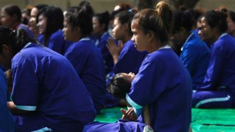 A woman in blue prison outfit cradling a baby sitting and looking forward.