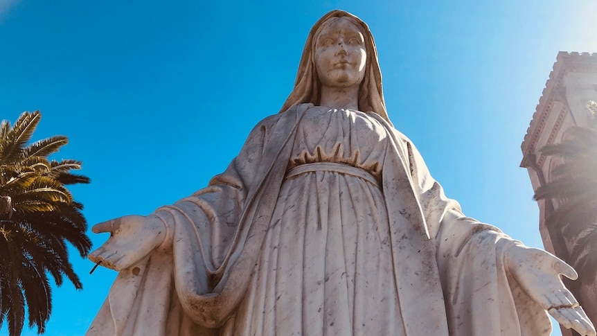 A statue of Mother Mary stands in front of a blue sky and church buildings