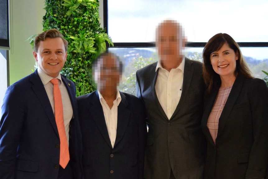 four people, two whose faces are blurred stand in front of a window in business attire