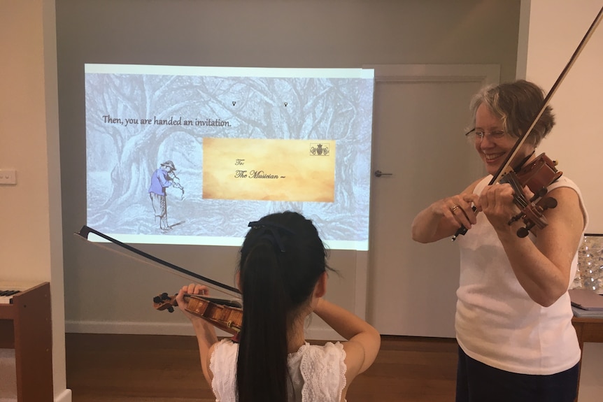 Sue playing the violin with her student (whose face isn't visible) as they look at a projector screen of the game