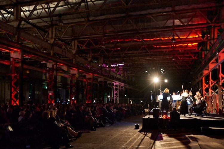 An industrial-style workshop with a stage and audience, with red lighting and an orchestra performing.