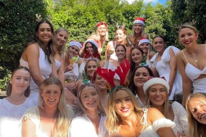 Un groupe de femmes, toutes vêtues de blanc, regarde la caméra, avec de grands arbustes en toile de fond