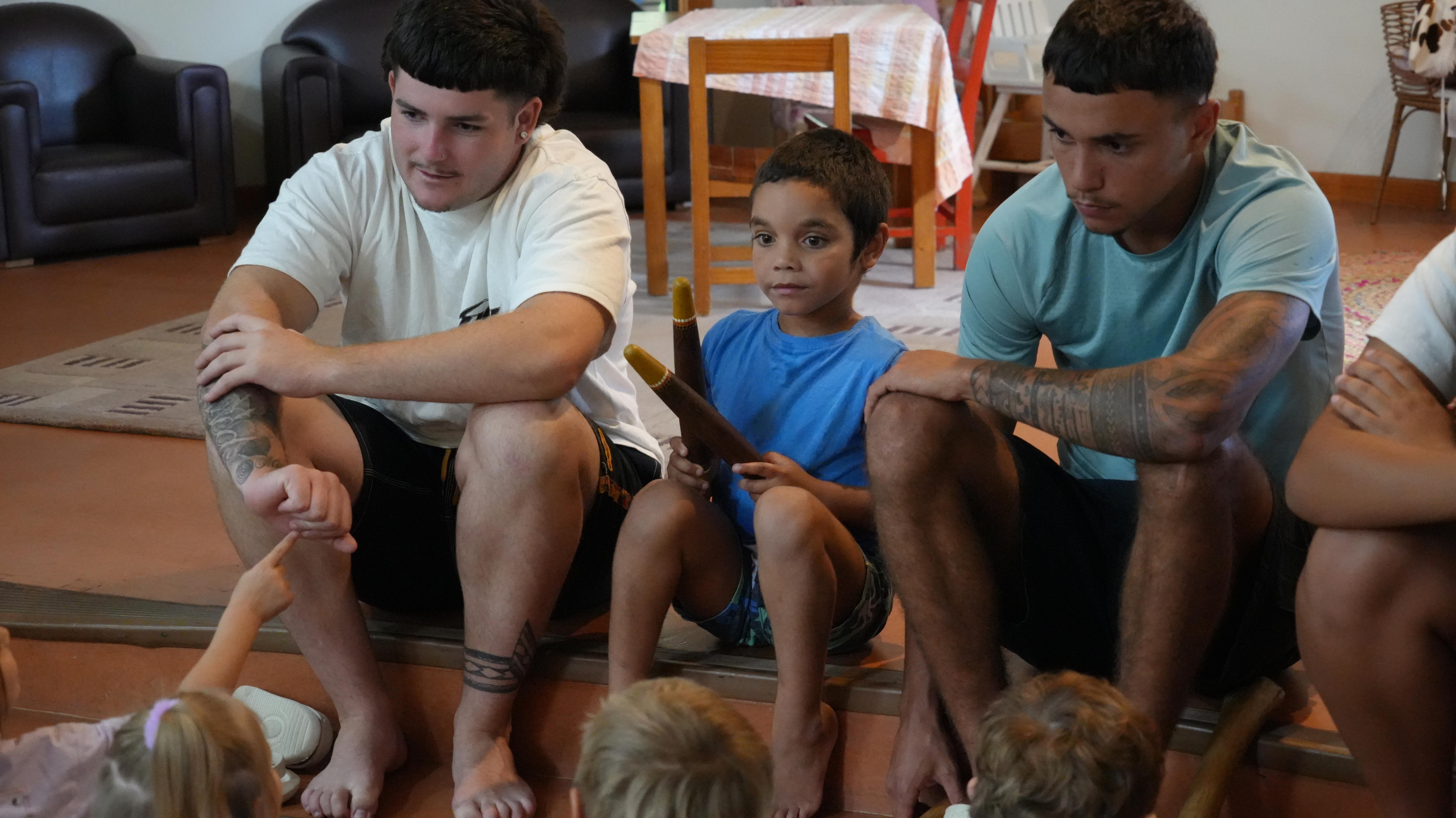 An Aboriginal boy sits with clapsticks between two young Aboriginal men sitting on a step in a learning space