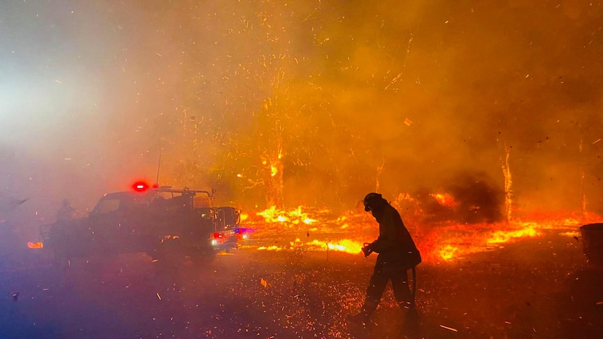 A firefigter moves through a fiercely burning fire ground at night, silhouetted by the fire.