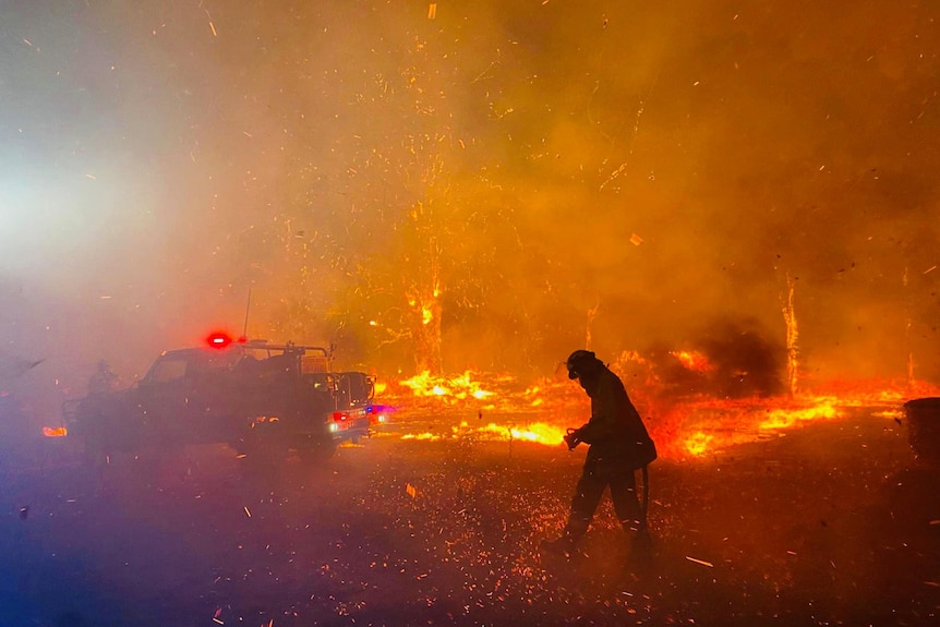 A firefigter moves through a fiercely burning fire ground at night, silhouetted by the fire.