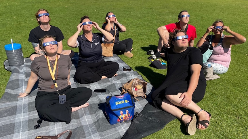 Seven people sit on a picnic blanket and look to the sky while wearing protective sunglasses. 