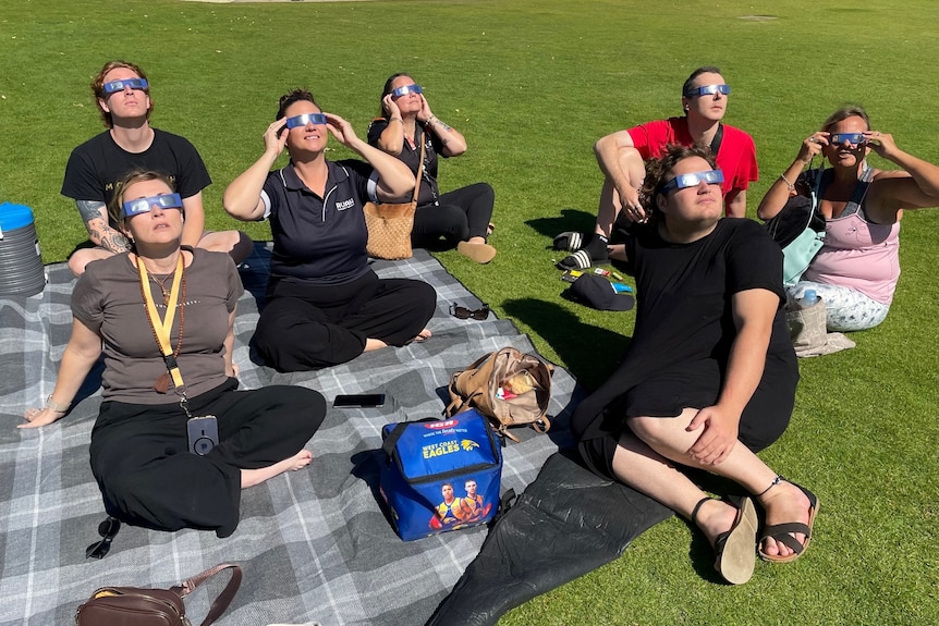 Seven people sit on a picnic blanket and look to the sky while wearing protective sunglasses.