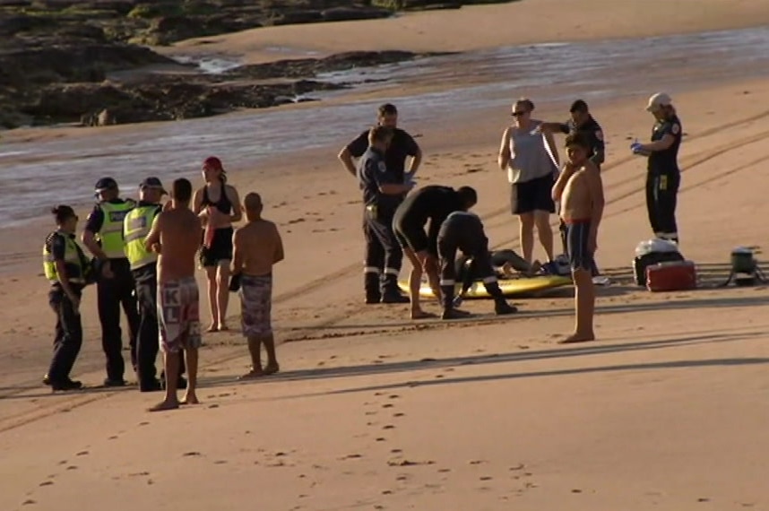 Volunteers and police stand on the beach near and around a man lying on a board.