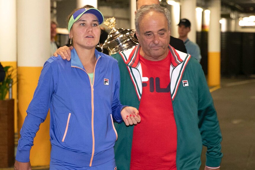 A female tennis player and her father walk ahead of a man holding the Australian Open women's trophy.