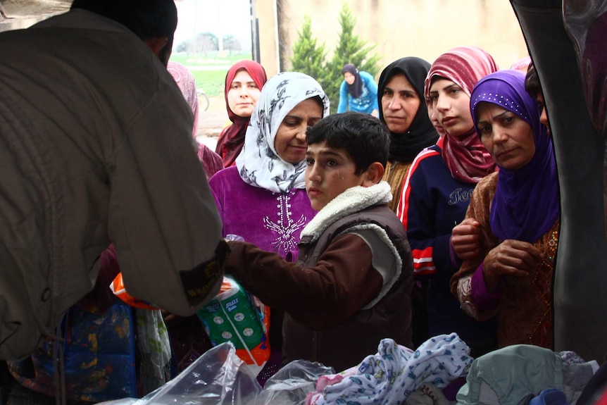A man hands out donations of clothes and goods to women and children from the back of a van.