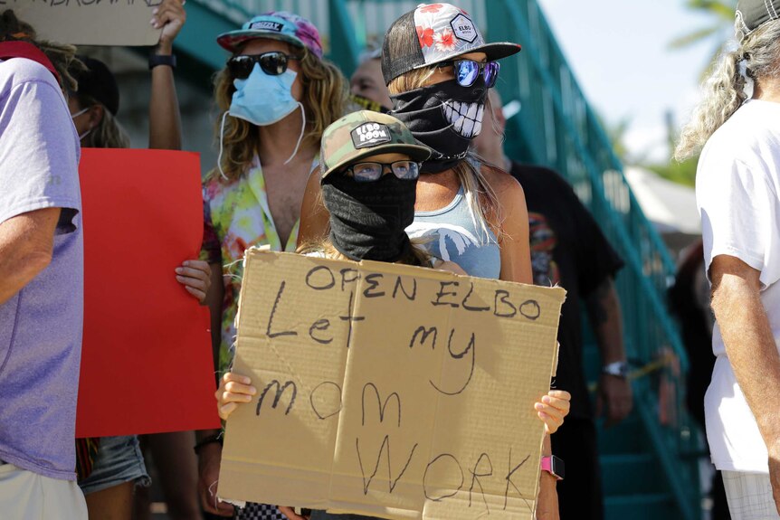 A masked child at a protest holds a sign saying Let My Mom Work