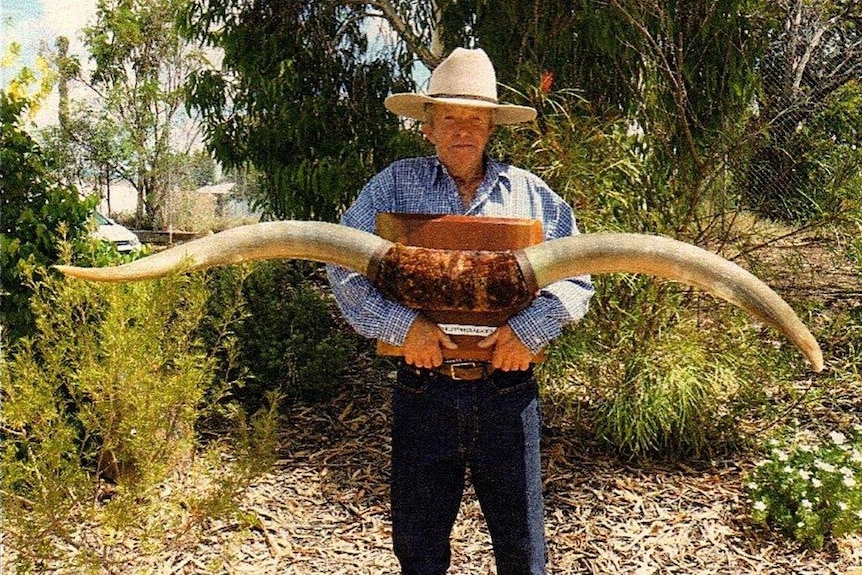 Man in Akubra hat holding a set of large bullock horns mounted on a timber plaque.