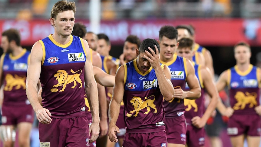 Dejected Brisbane Lions players walk off the Gabba, with one rubbing his head with his hand.