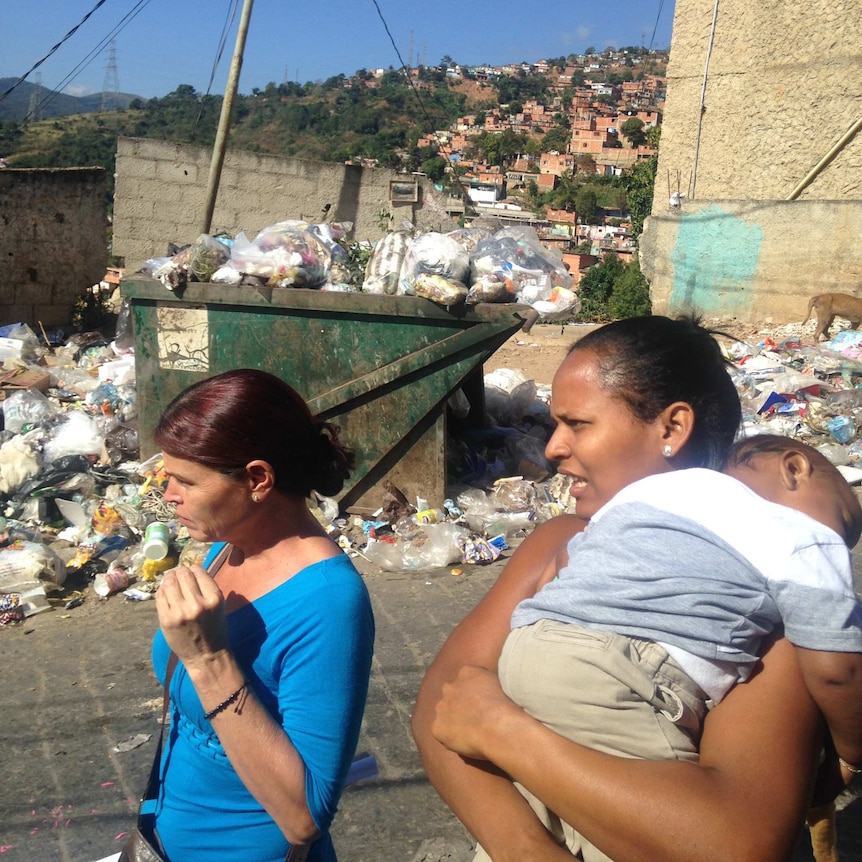 People walk past an overflowing garbage skip with a barrio stretched over a hillside in the background.