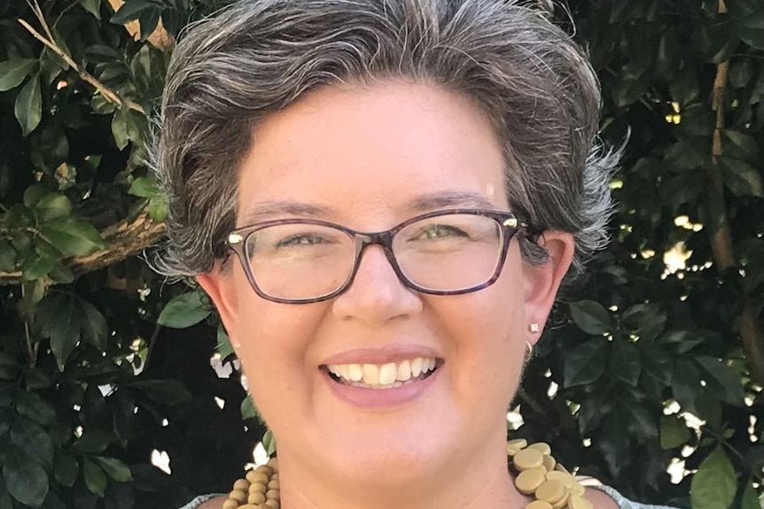 A woman with grey hair and glasses smiles. She is wearing a green shirt and a necklace. There is foliage in the background.