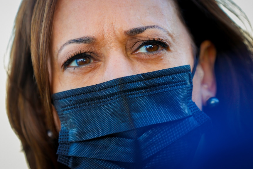 A close up of Kamala Harris, wearing a black surgical mask and pearl earrings