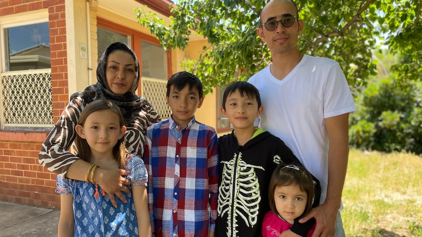 A woman and a man stand with four young children between them. Behind them is a brick house