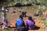 The Country Hour's Matt Brann interviews Selma Thompson on a creek bank while children play in the water