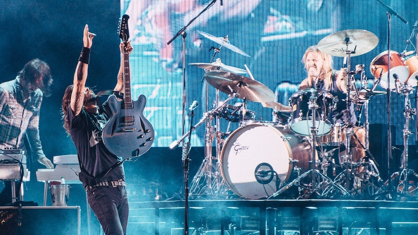 Dave Grohl of Foo Fighters raises his arm and guitar in the air on stage. Taylor Hawkins plays drums and grins