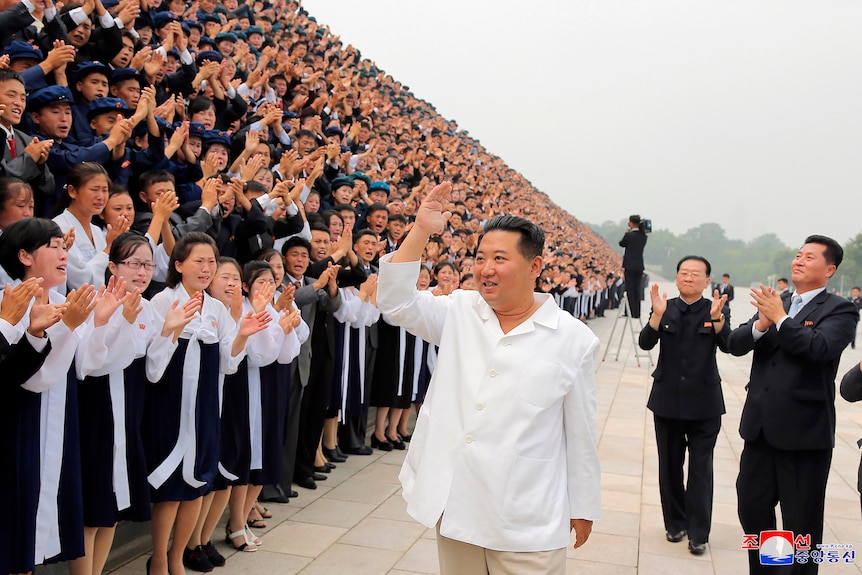 Kim Jong Un wears a white shirt as he stands waving to a crowd of people clapping.