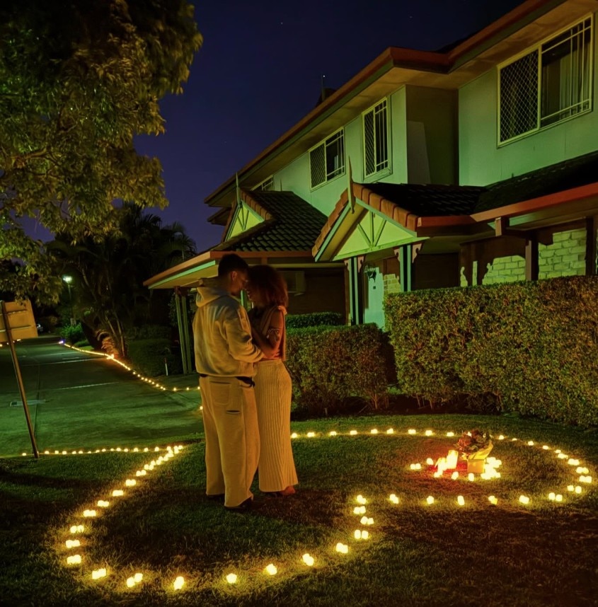 A young man and a young woman embrace on a lawn, which is surrounded by tealight candles in a love heart shape.