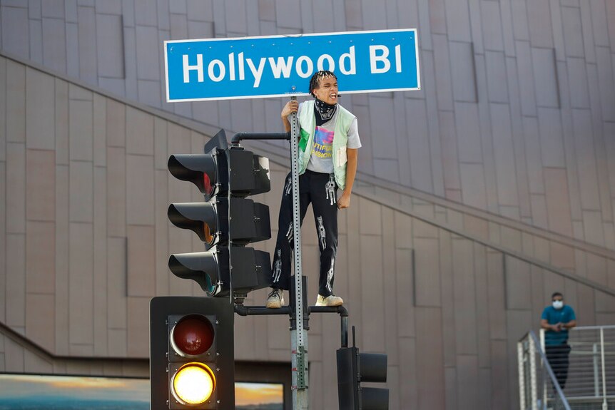 You view a man standing atop traffic lights as he holds onto a street sign that reads 'Hollywood Bl'.