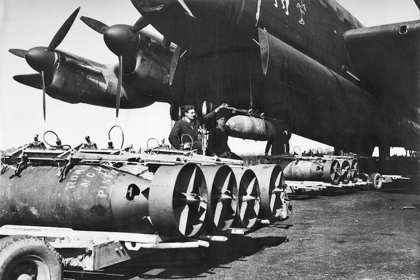 A plane on an airfield with worker loading bombs into the bottom of the aircraft.