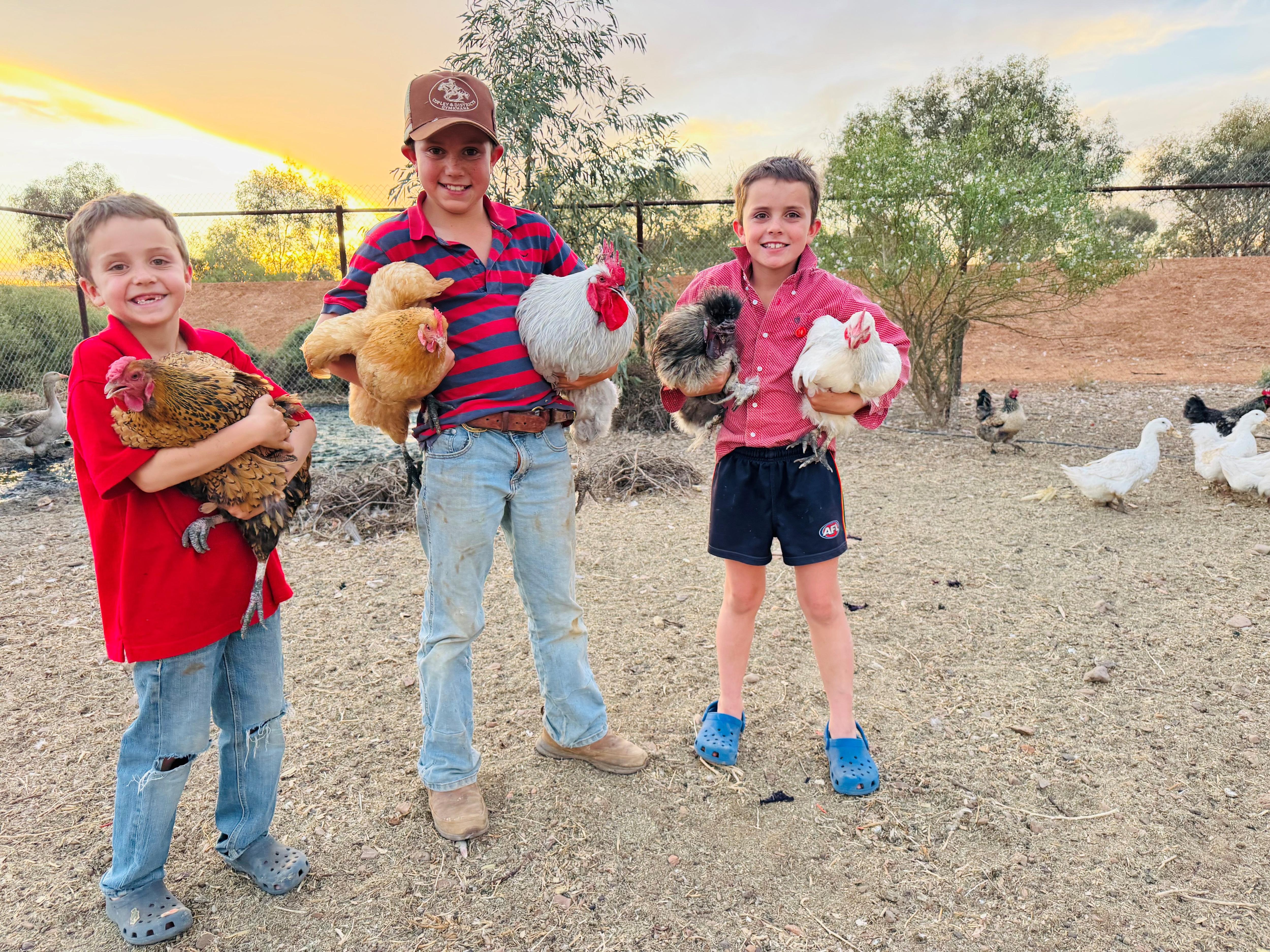 Tres niños se paran frente a una puesta de sol, cada uno con un gancho en la mano y mirando a la cámara.