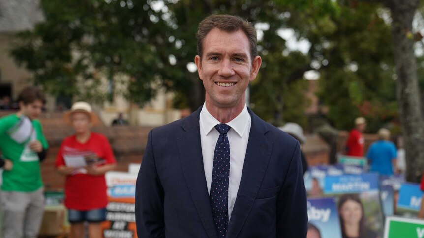 Sam Crosby stands smiling, looking at the camera, in front of campaign posters and party volunteers.